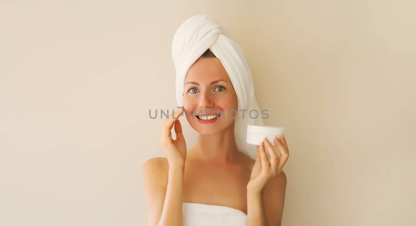 Natural beauty portrait of happy smiling young caucasian woman touches her clean skin applying face cream while drying wet hair with white bath towel on her head