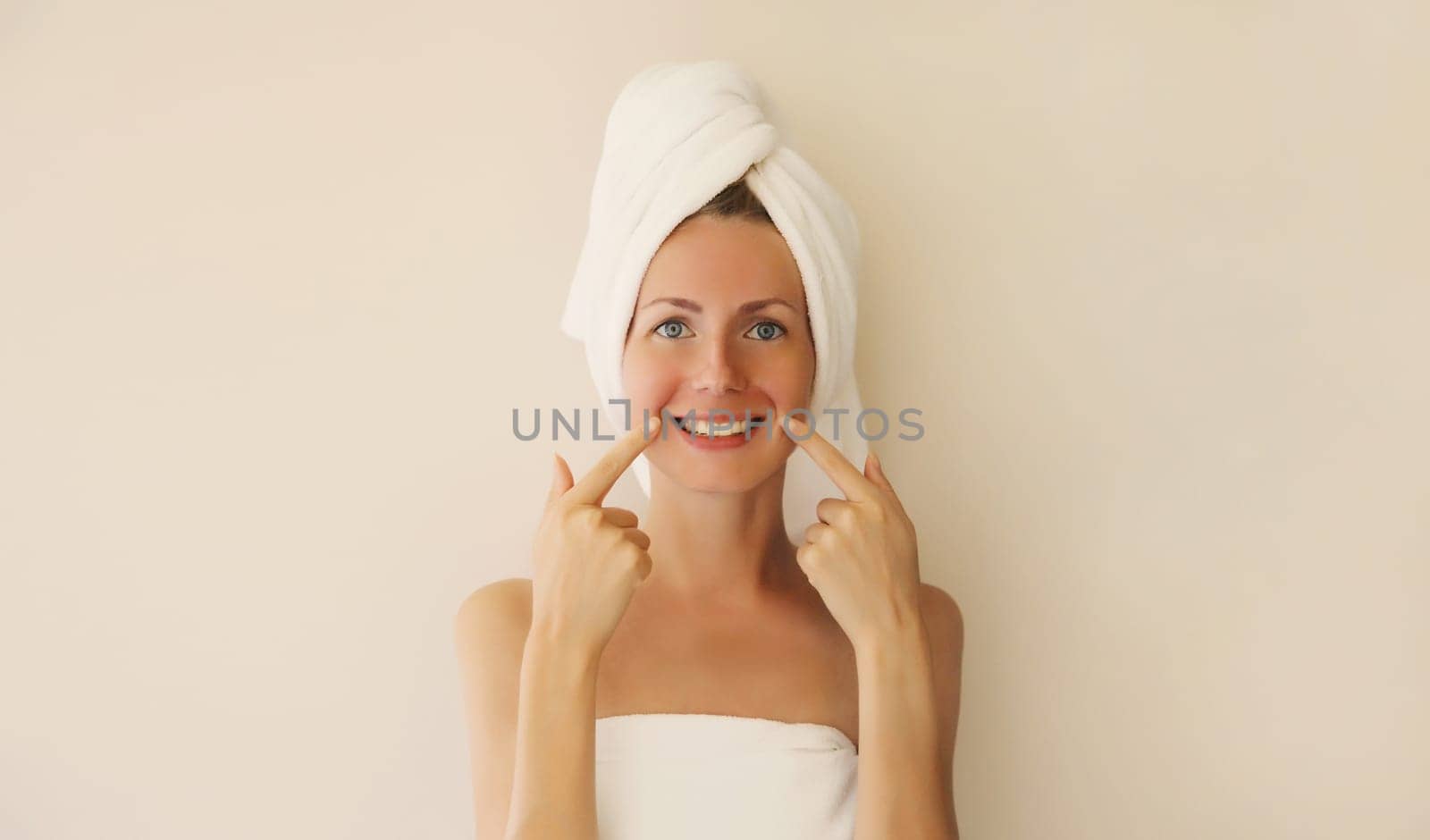 Portrait of happy smiling caucasian young woman points to her white clean teeth while drying her hair with towel on her head after shower in the morning at home