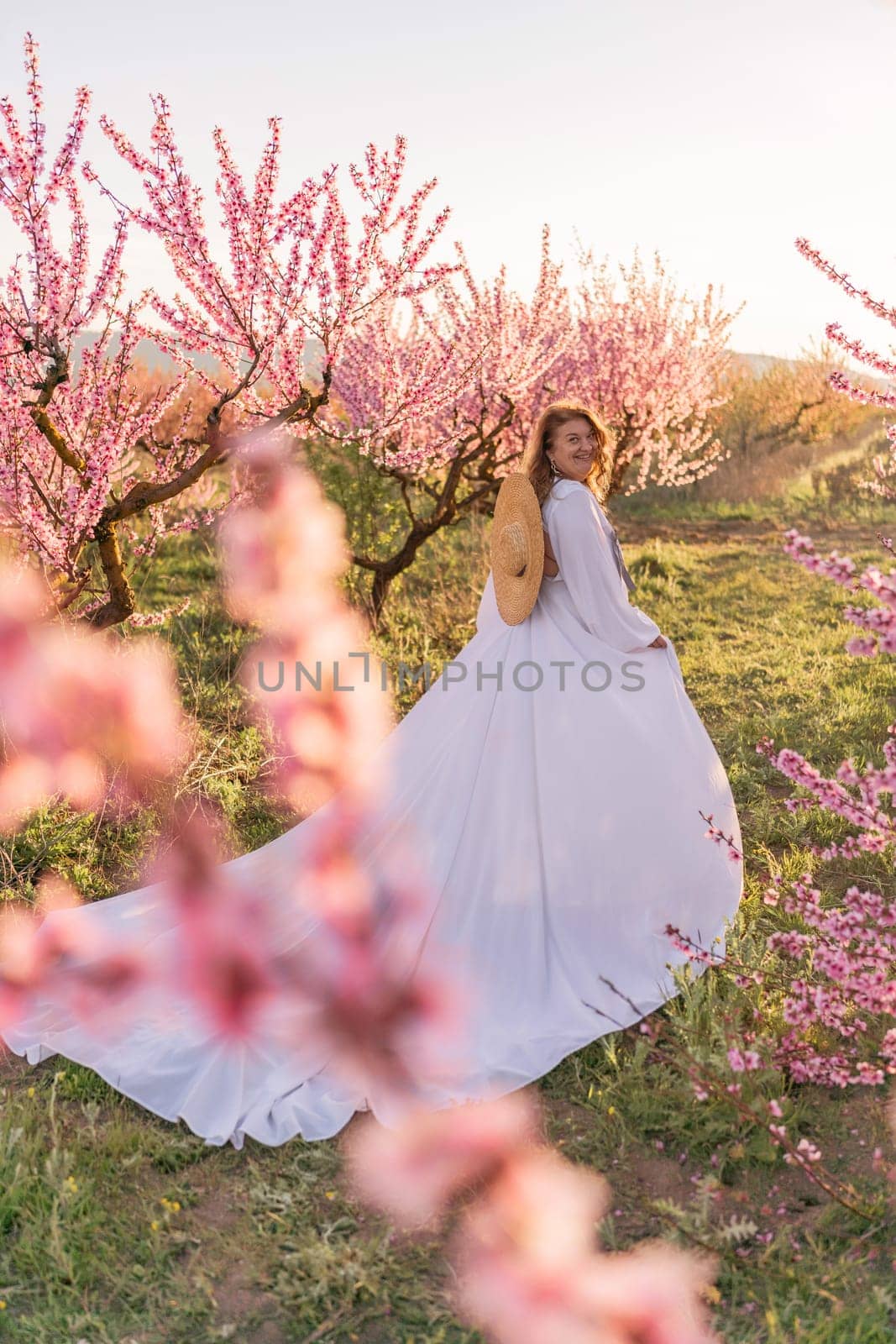 Woman blooming peach orchard. Against the backdrop of a picturesque peach orchard, a woman in a long white dress and hat enjoys a peaceful walk in the park, surrounded by the beauty of nature
