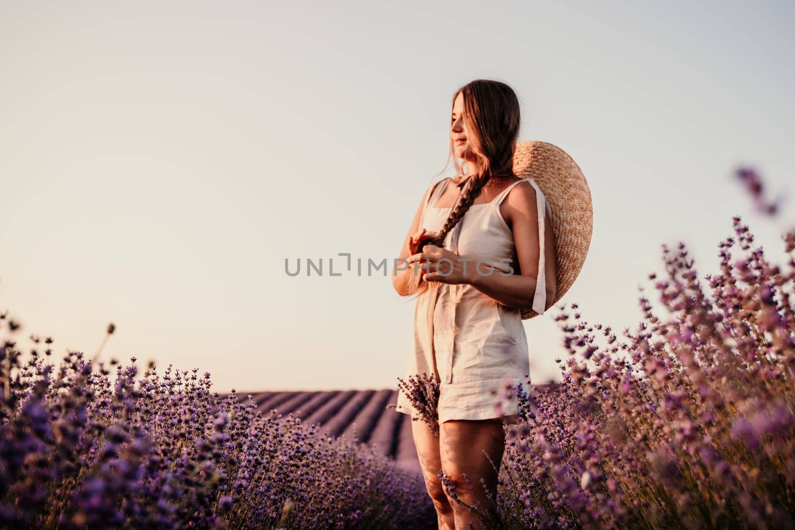 A woman is walking through a field of purple flowers with a straw hat on.