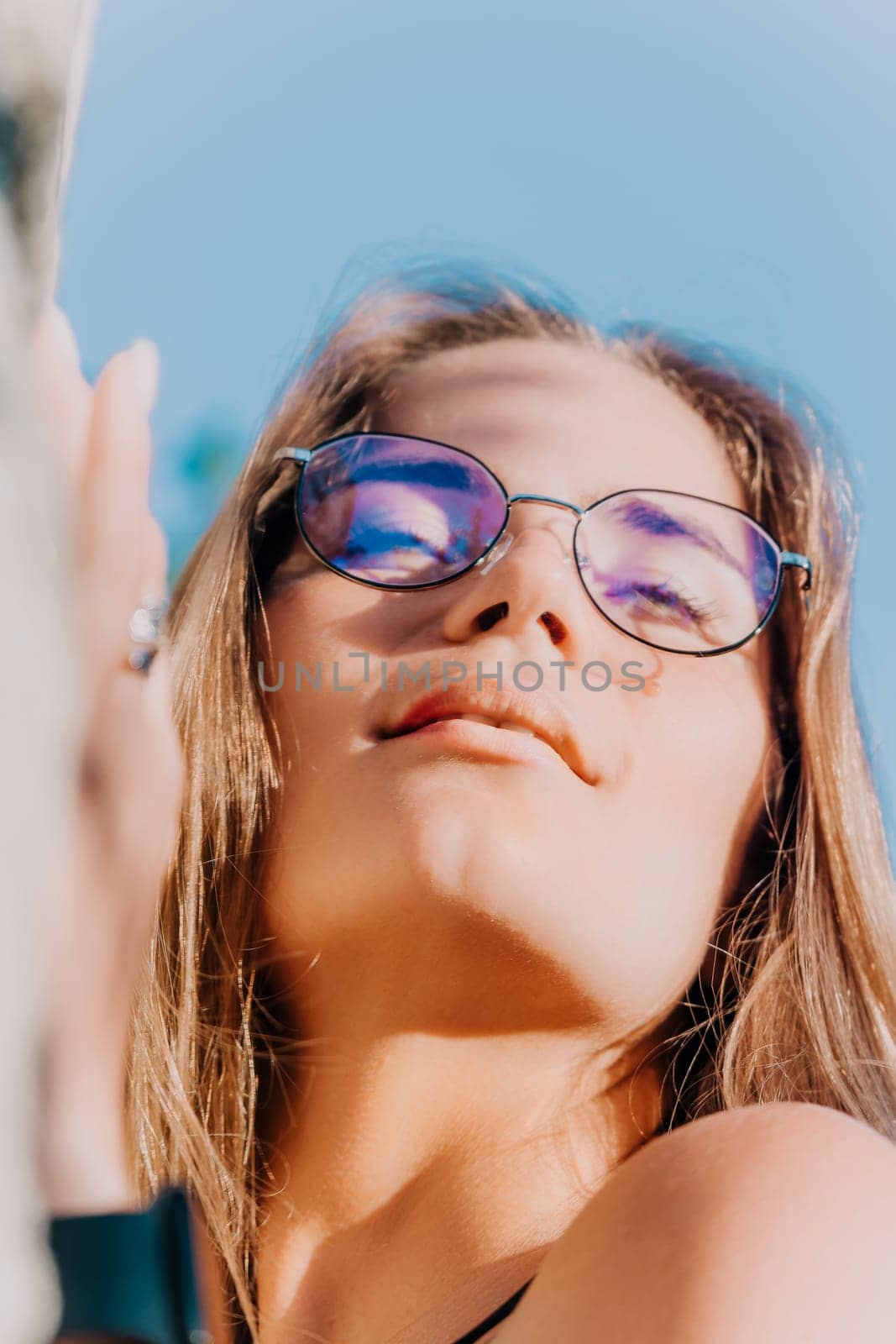 A woman with long blonde hair wearing purple-tinted glasses looks up at the sun.
