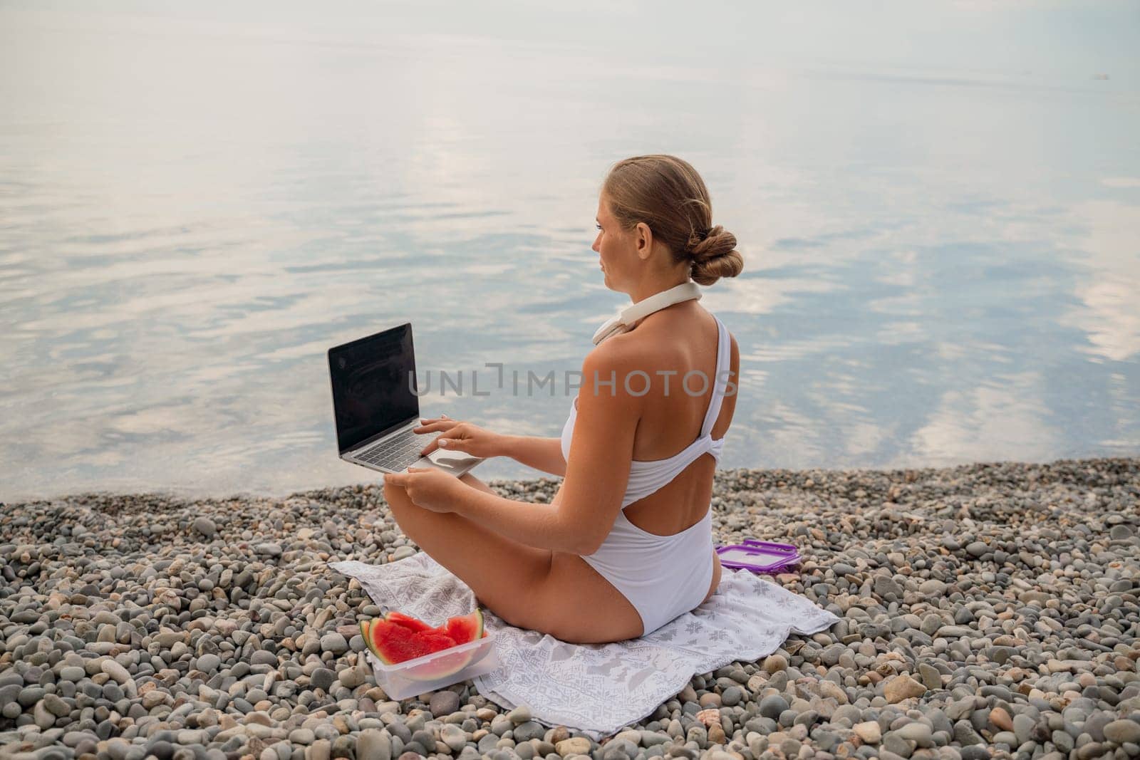 A woman is sitting on the beach with a laptop open in front of her. She is wearing a white bikini and has a pink headband on. Concept of relaxation and leisure