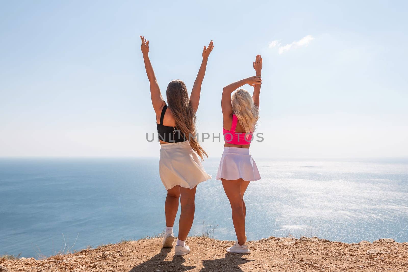 Two women are standing on a beach, one wearing a pink top and the other wearing a black top. They are both smiling and raising their arms in the air, as if they are celebrating something