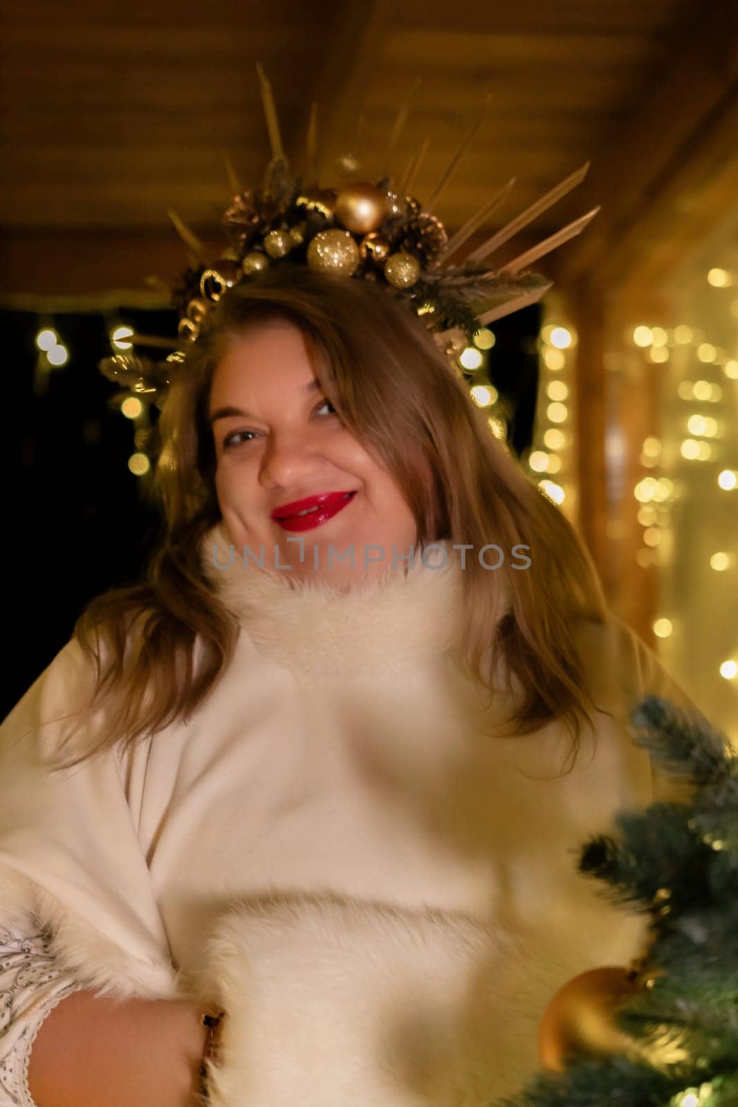A woman with a festive crown of ornaments and branches smiles warmly. String lights twinkle in the background, suggesting a Christmas setting.