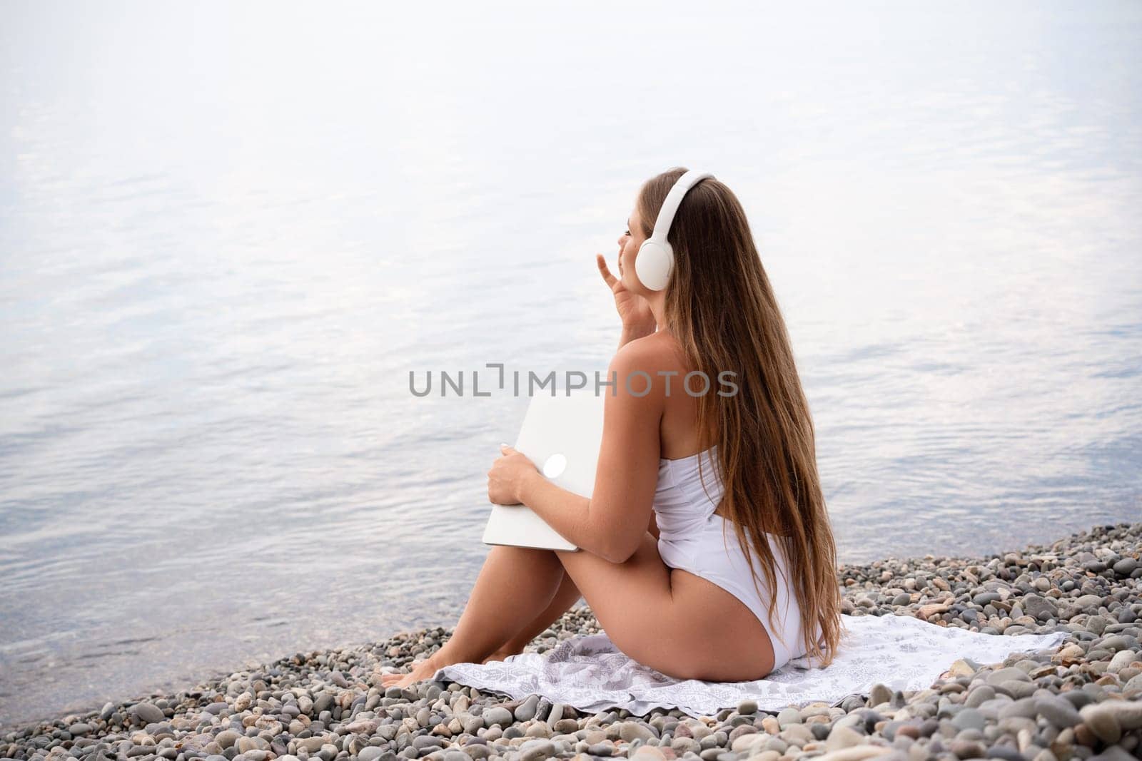 A woman is sitting on the beach with a laptop and headphones on. She is wearing a white bikini and she is enjoying her time by the water