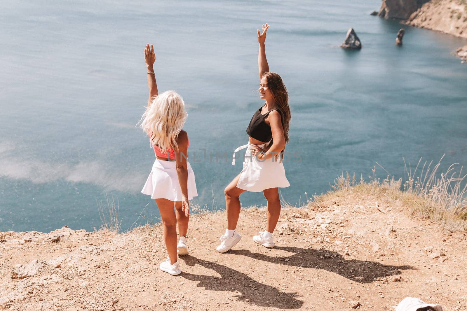 Two women are standing on a hill overlooking the ocean, one wearing a pink top and the other wearing a black top. They are both smiling and seem to be enjoying the view