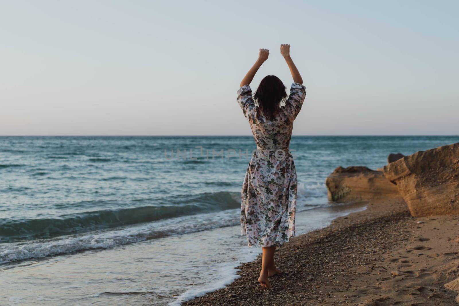 A woman in a floral dress stands on a sandy beach, arms raised in the air, facing the ocean. The water is choppy and the sky is blue.