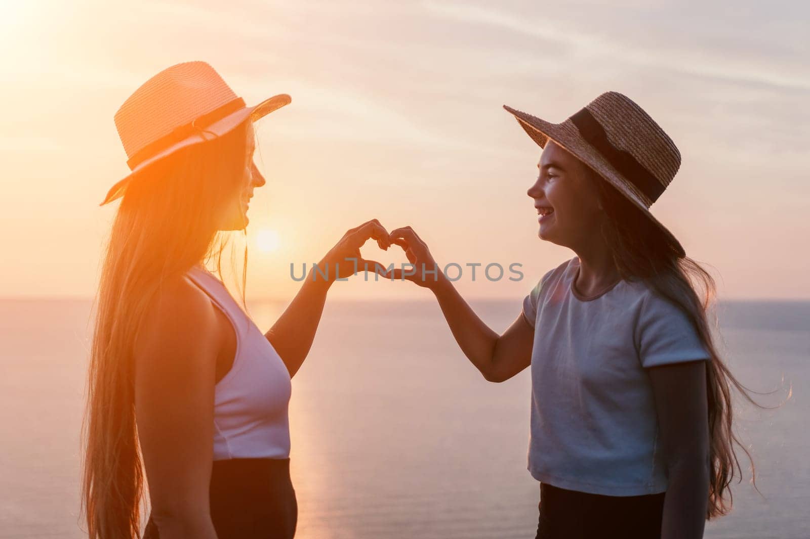Two women wearing straw hats stand side by side, facing each other and making a heart shape with their hands as the sun sets over the ocean.
