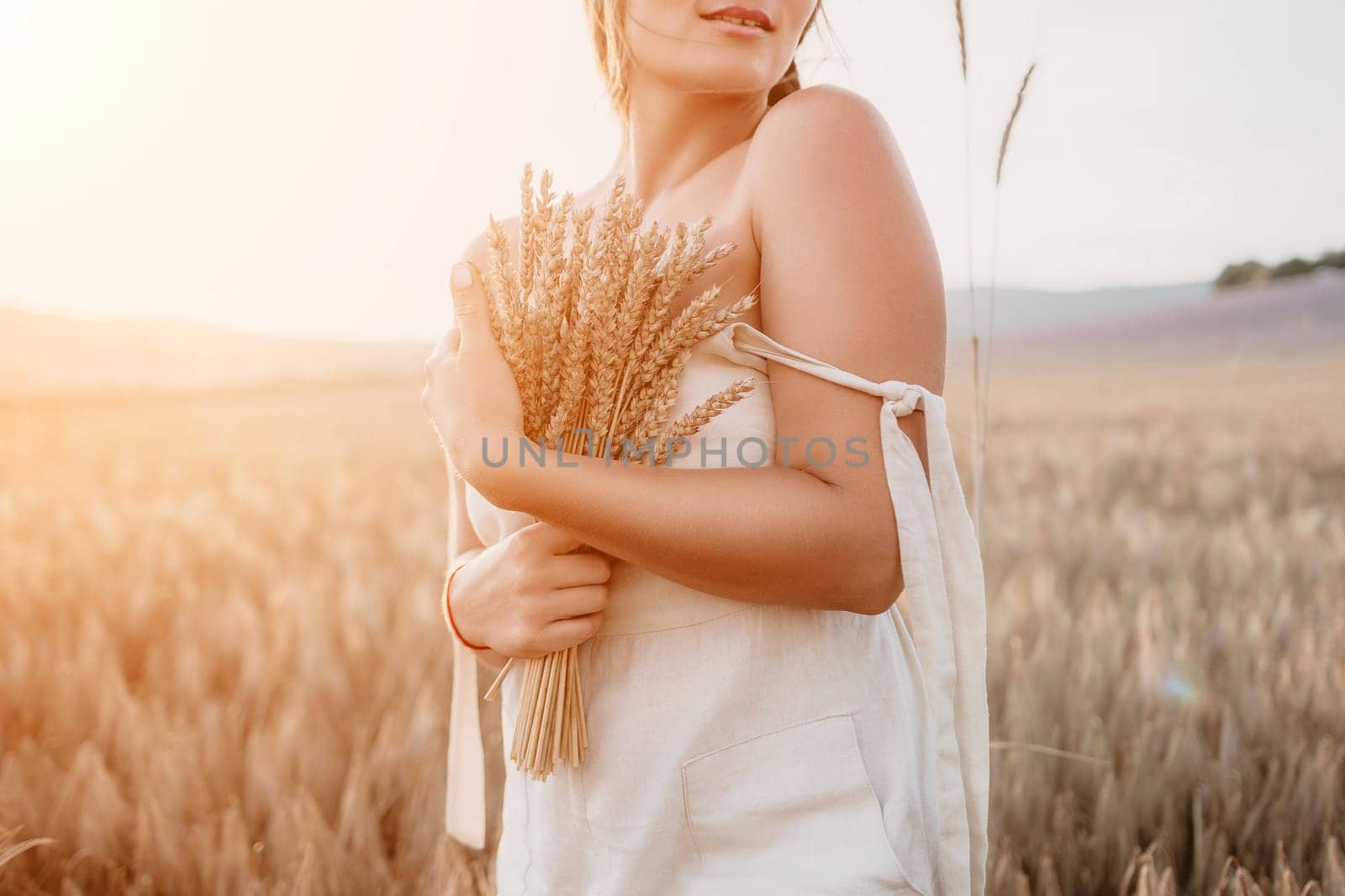 A woman is holding a bunch of wheat in her arms. The wheat is dry and brown, and the woman is wearing a white dress. The scene is set in a field, and the woman is posing for a photo