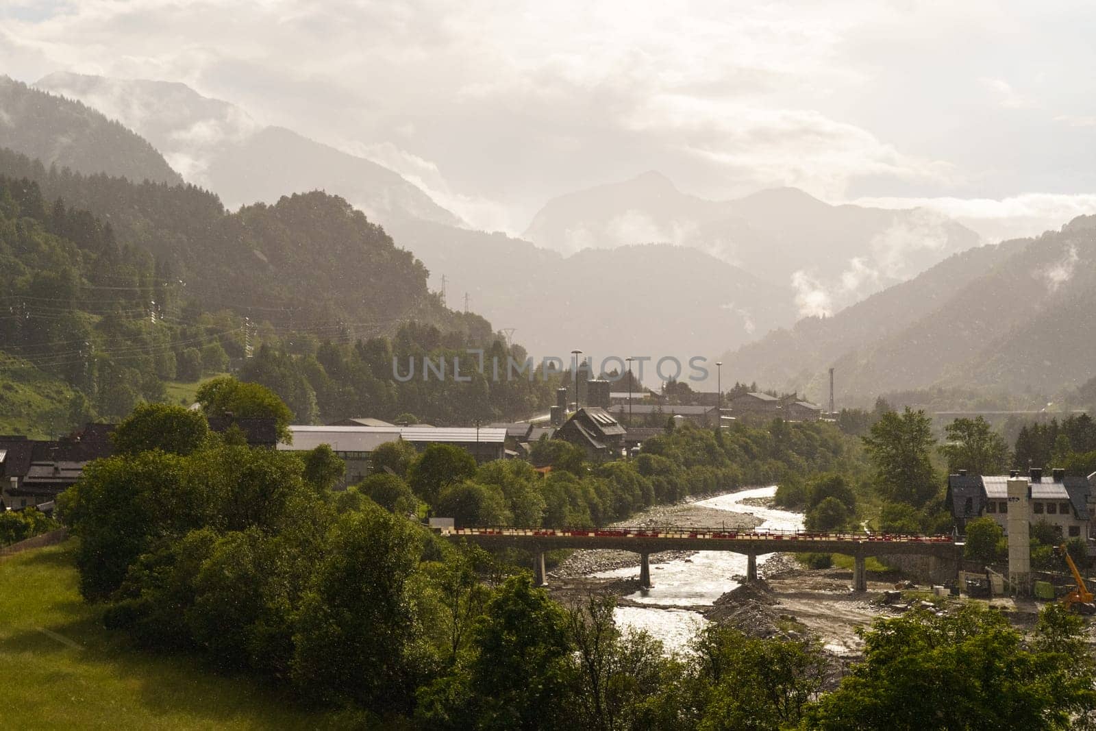 The sun rises over a tranquil valley, illuminating the river winding through vibrant green trees. Houses peek through the landscape as distant mountains fade into the mist.