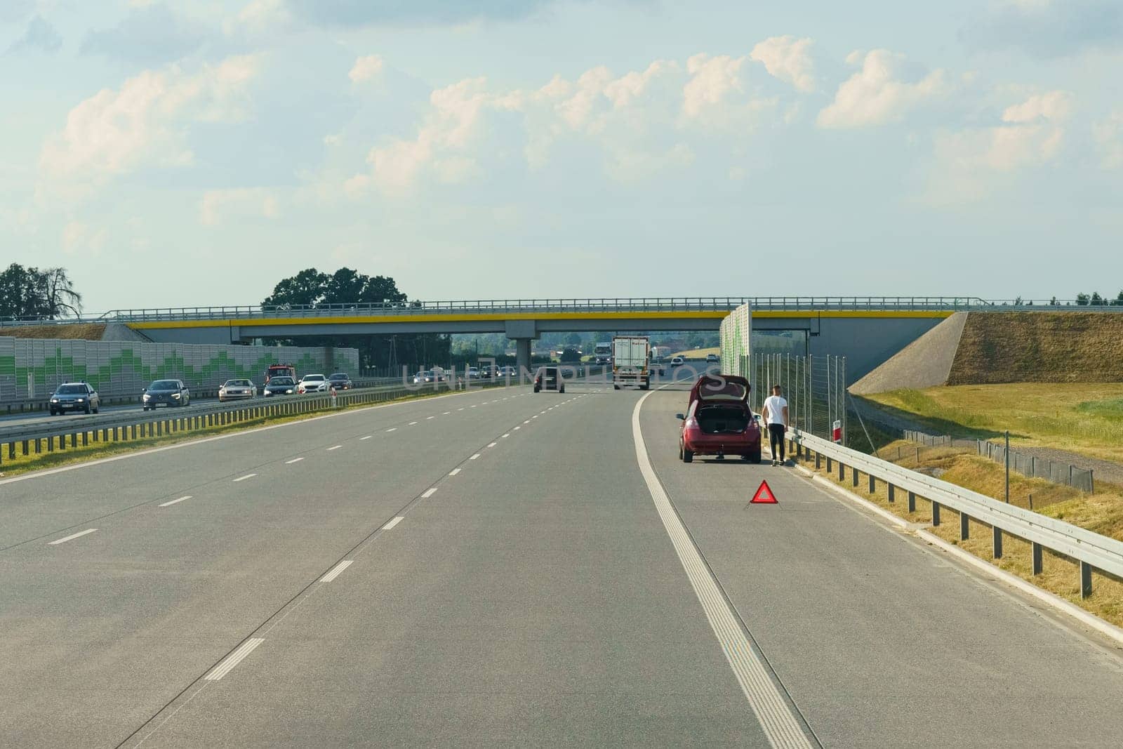 Gliwice, Poland - June 15, 2023: A man stands beside a red car with its trunk open on the side of a highway, placing a warning triangle on the road. The landscape features fields under a clear blue sky.