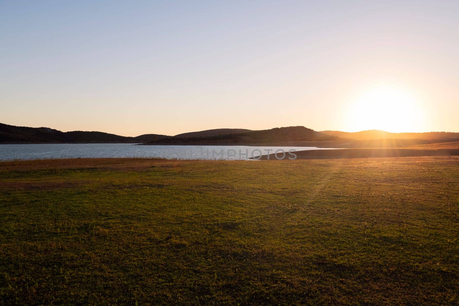 the sun in the clear sky is setting casting a warm glow over the land with a lake reflecting the sunlight, concept of peaceful and serene landscape, copyspace for text