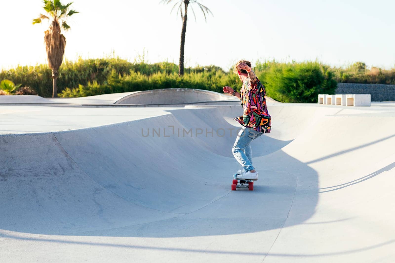 back view of anonymous funky young male skateboarder in trendy colorful shirt and jeans performing trick on concrete ramp while practicing skills in skatepark