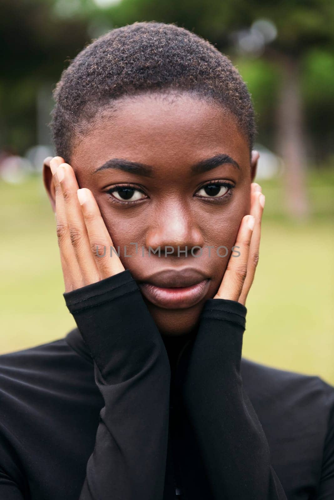 young tender african american female with short hair covering face with hands while looking at camera outdoors on summer day