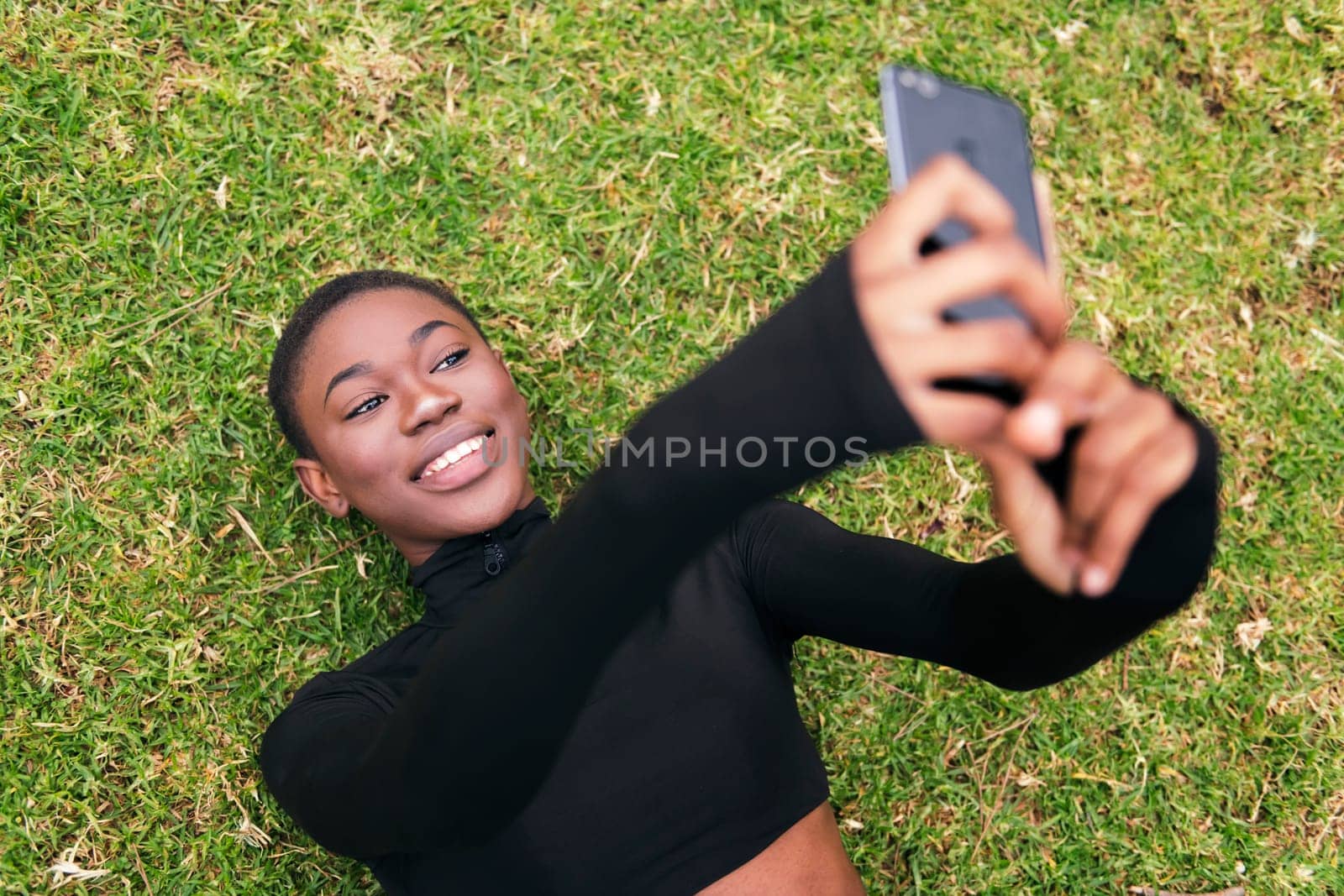 young african american woman with short hair smiling happy taking selfie photo on mobile phone lying on the grass in a park