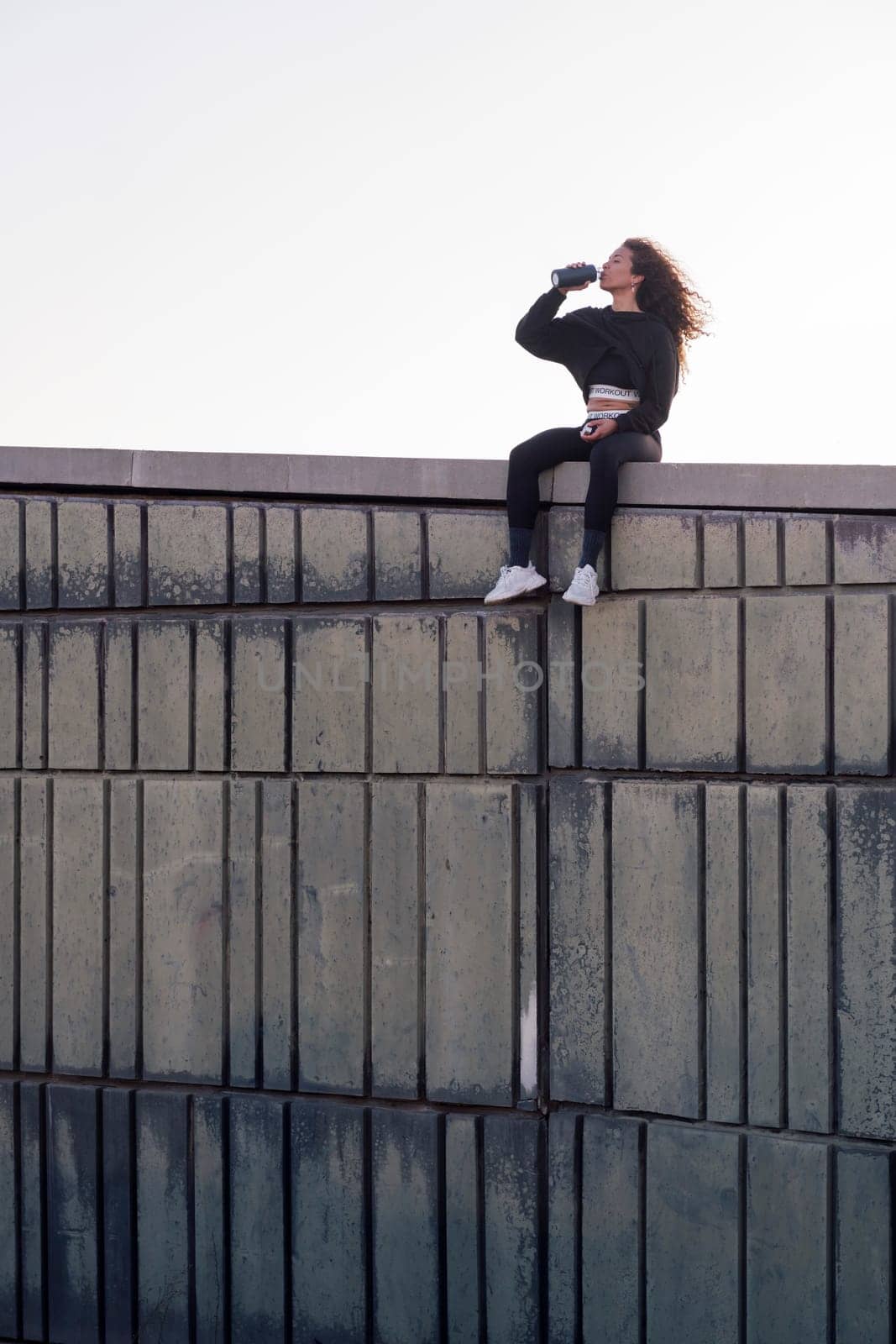 thirsty female athlete in sports clothes drinking beverage from bottle while resting on building roof after workout, concept of urban sport and healthy lifestyle