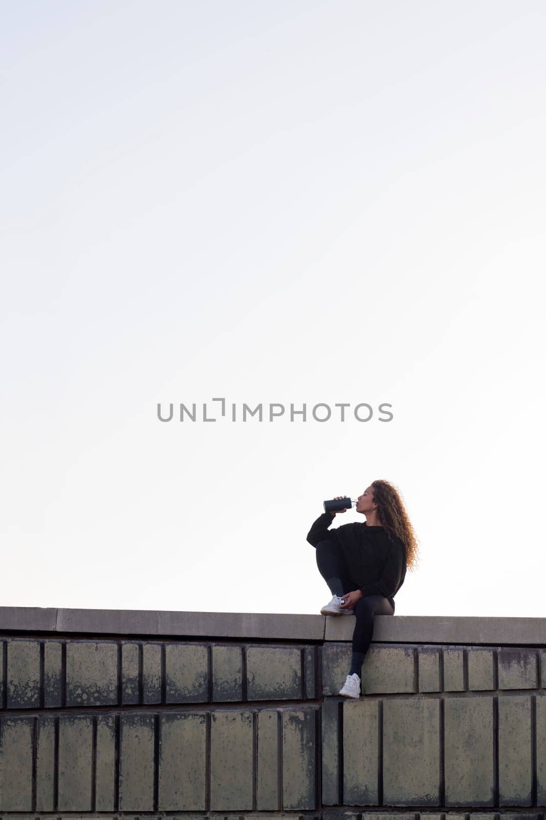 thirsty female athlete in sports clothes drinking beverage from bottle while resting on building roof under light sky, concept of urban sports and healthy lifestyle