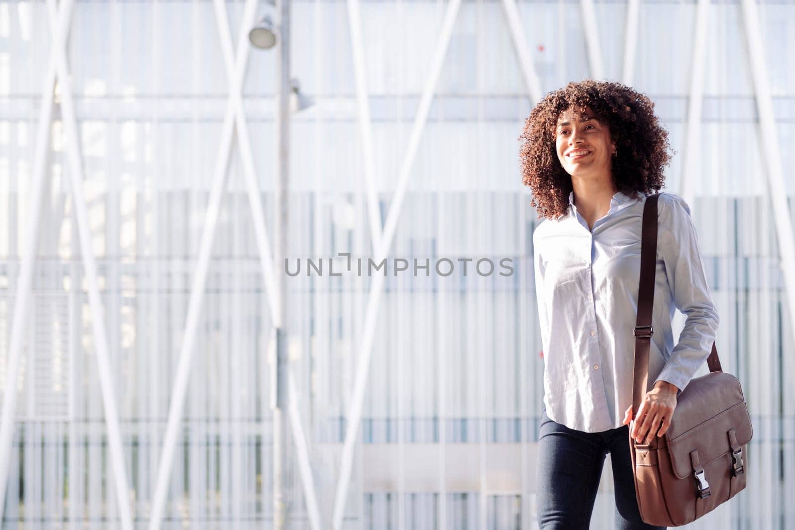 smiling female entrepreneur in smart casual outfit walking along street in megalopolis and looking away