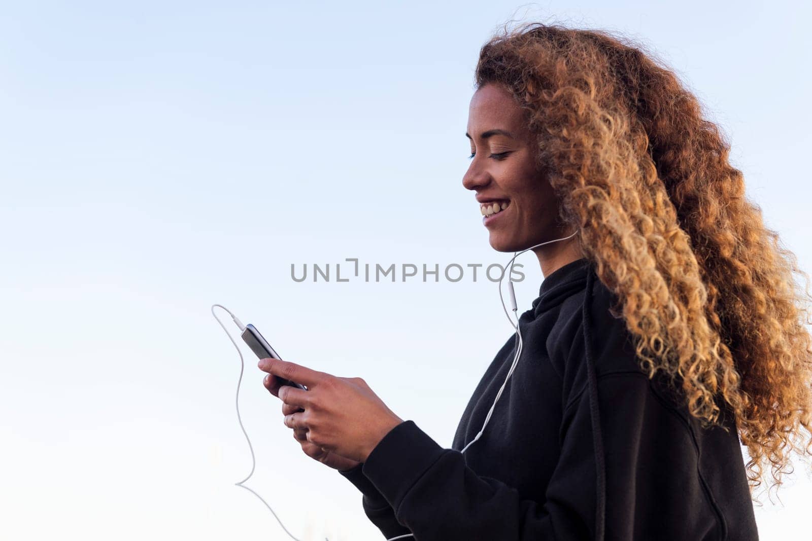 latin female athlete in sports clothes smiling happy while listening music on earphones from cellphone, concept of urban sport and healthy lifestyle