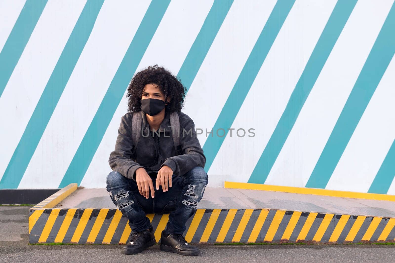 serious curly haired african american male teenager in black protective mask for flu prevention looking away thoughtfully sitting against striped wall on street