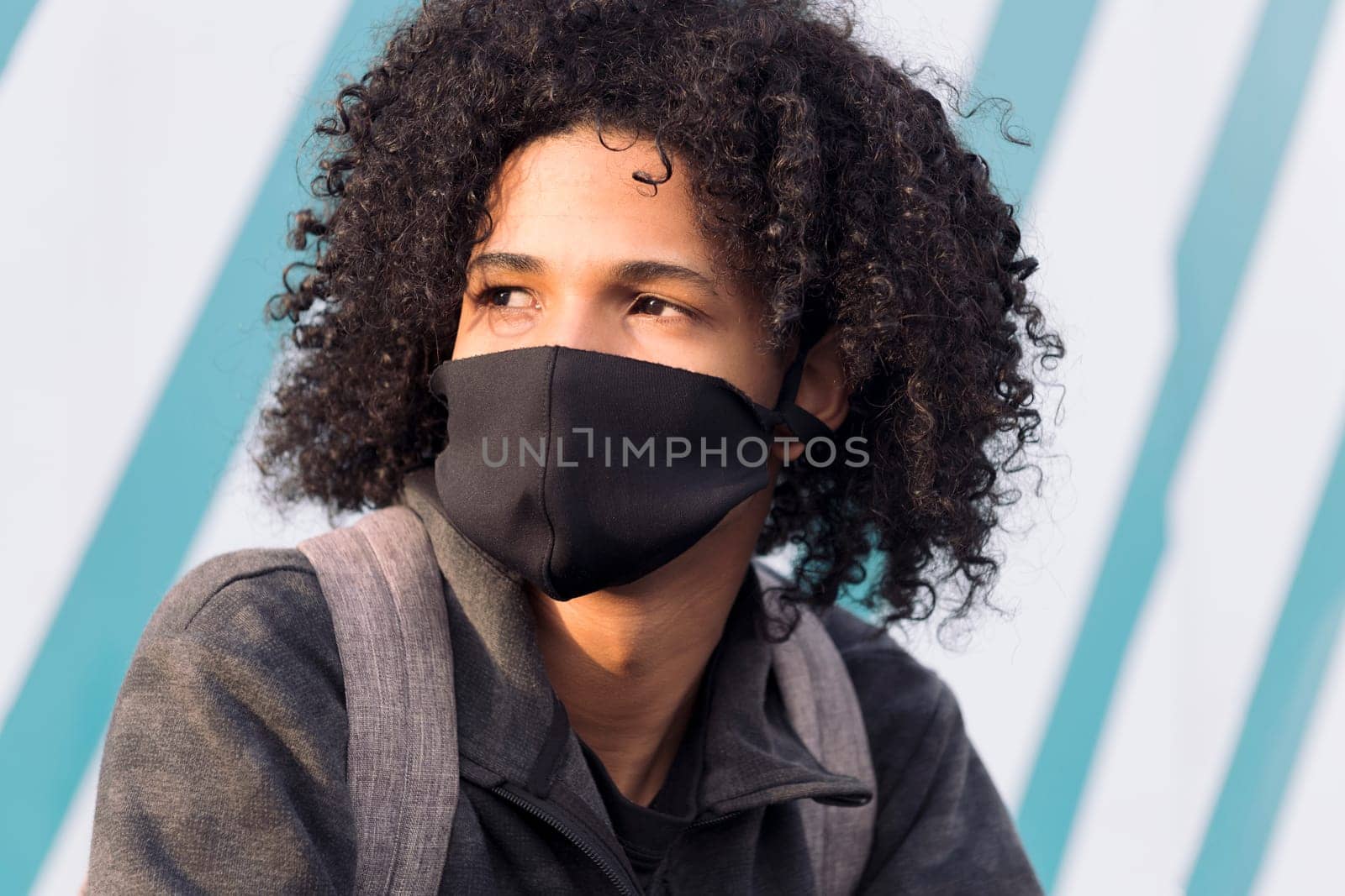 serious curly haired african american male teenager in black protective mask for flu prevention looking away thoughtfully against striped wall on street