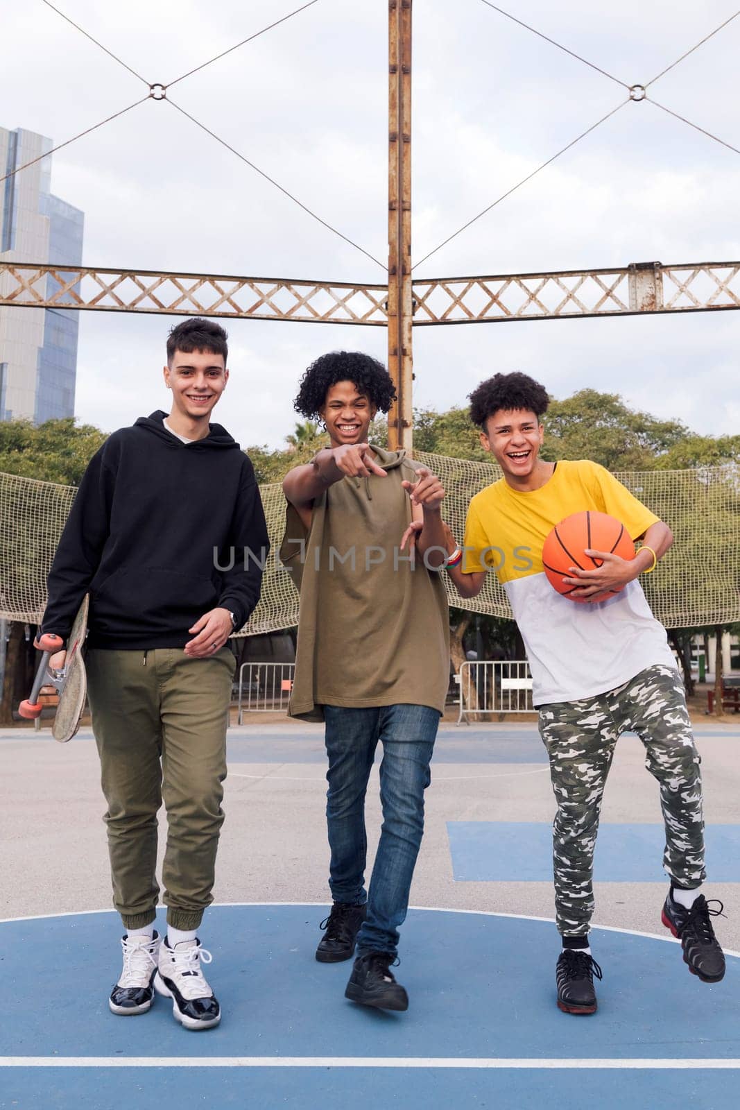 three confident multiracial male teens with basketball and skateboard walking joking and laughing happy while having fun in a sports court at park, concept of youth and urban lifestyle