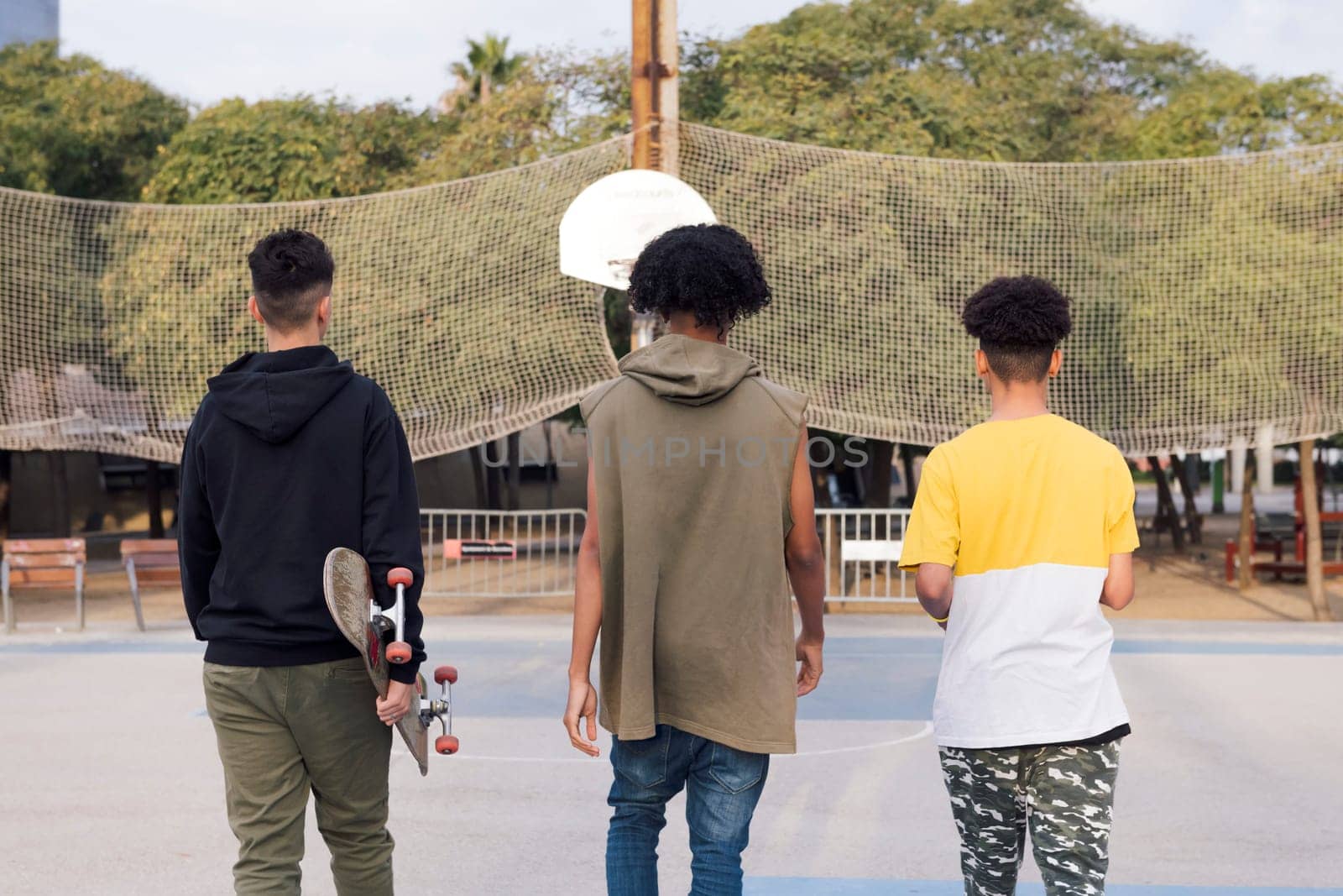 rear view of three unrecognizable multiracial male teens with skateboard walking in a sports court at park, concept of youth and urban lifestyle