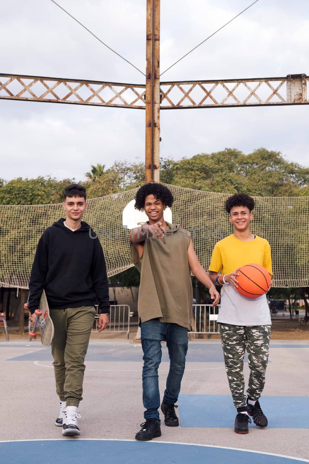 three confident multiracial male teens with basketball and skateboard walking joking and smiling happy while having fun in a sports court at park, concept of youth and urban lifestyle