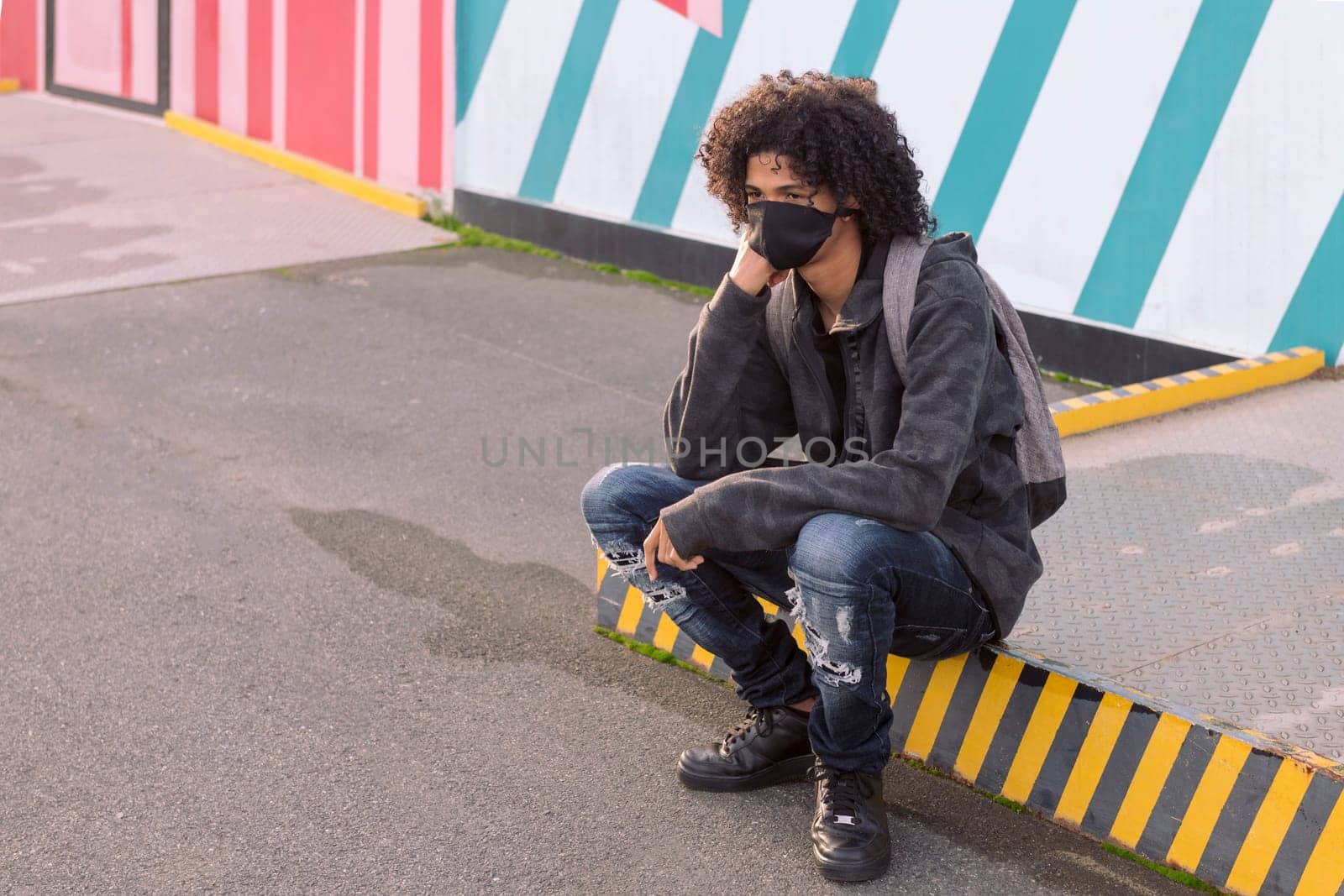 serious curly haired african american male teenager in black protective mask for flu prevention looking away thoughtfully sitting against striped wall on street