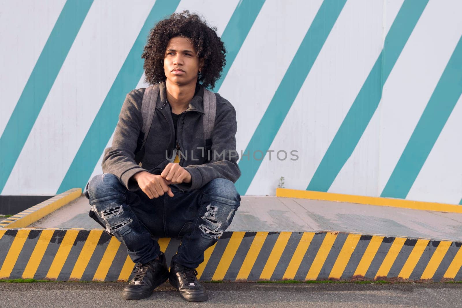 serious curly haired african american male teenager in informal street wear looking away thoughtfully sitting against striped wall on street