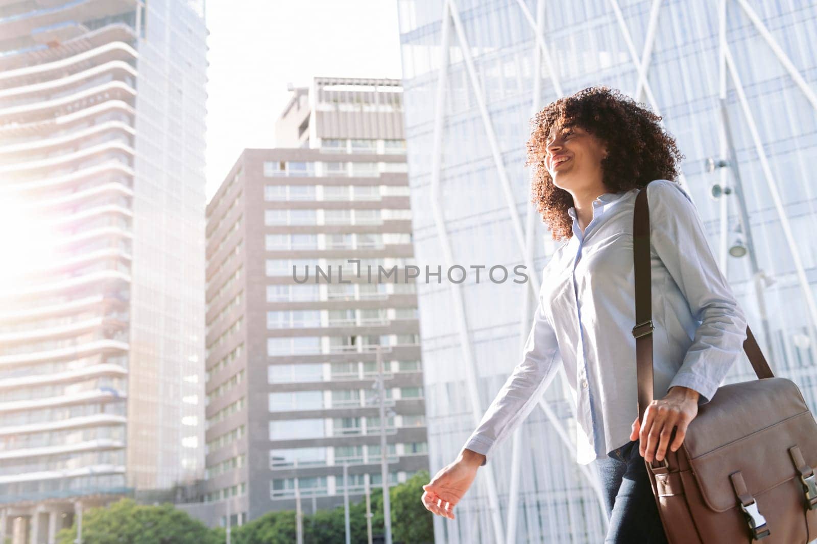 low angle side view of smiling female entrepreneur in smart casual outfit walking along street in city and looking away