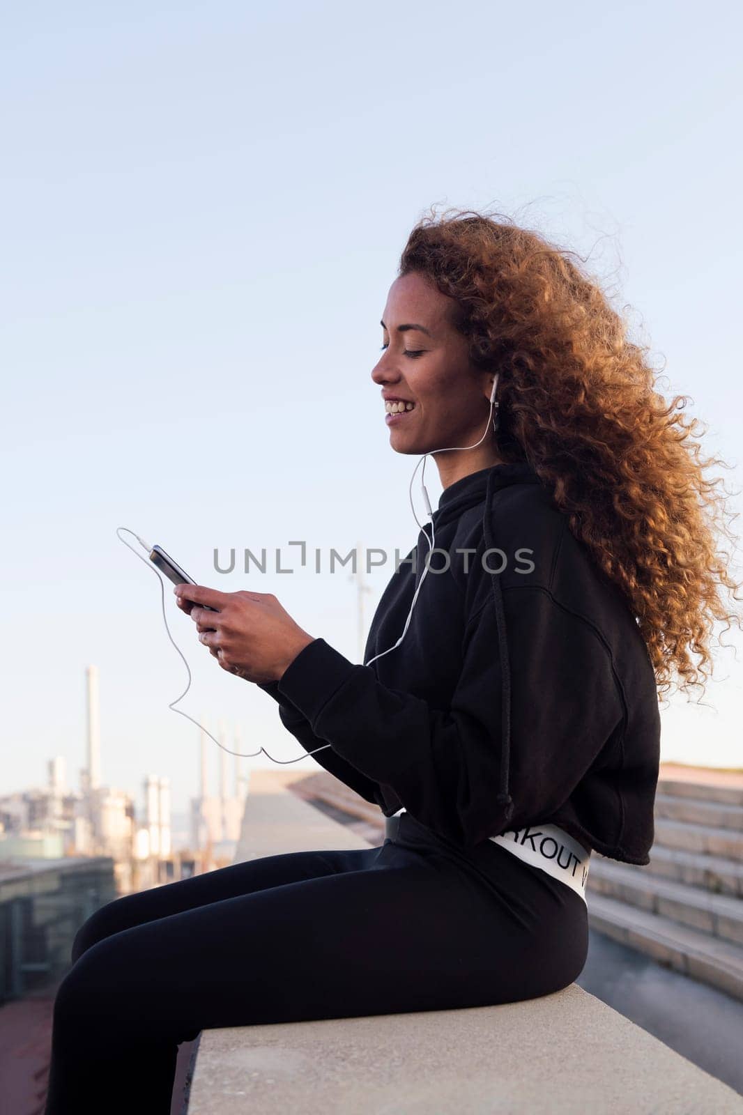 side view of content african american female athlete in earphones text messaging on cellphone while having break from workout in city, concept of urban sport and technology of communication