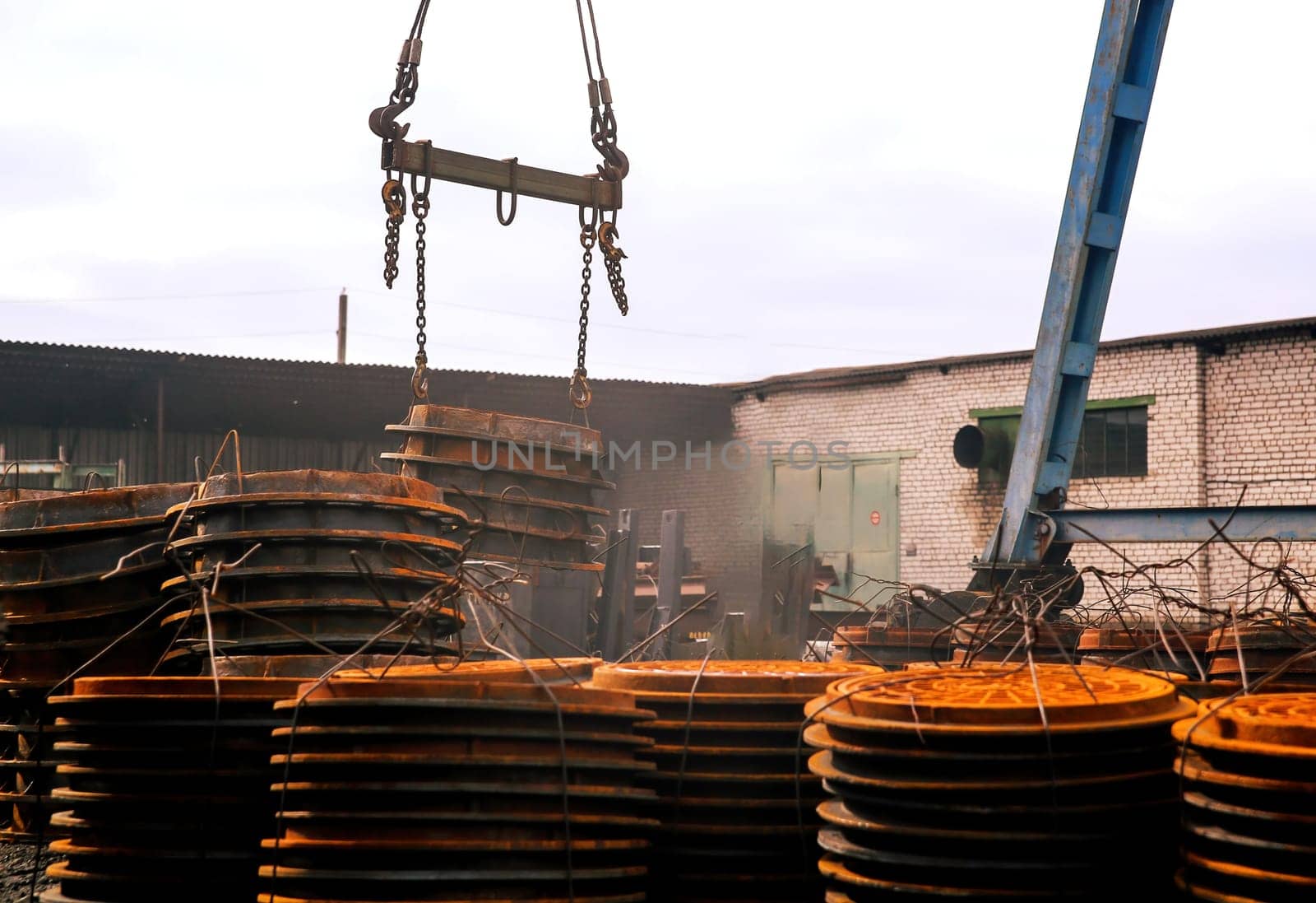 A crane hoists large, circular metal plates at an industrial site. Surrounding stacks of rusty plates indicate ongoing construction during overcast weather.