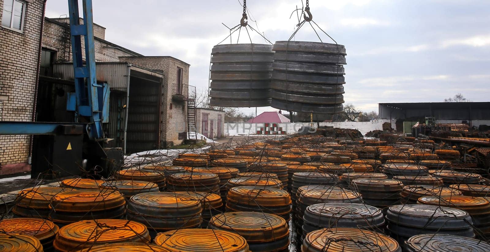 A crane is actively lifting heavy metal coils at a storage facility, surrounded by stacks of similar coils on a chilly, overcast day. The environment highlights industrial operations.