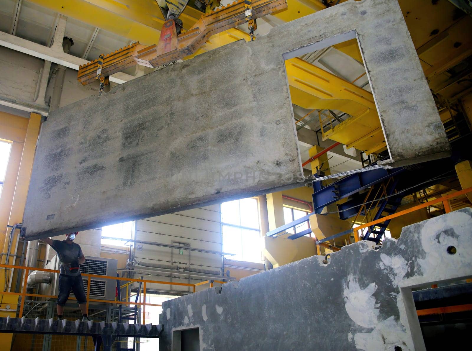 A construction worker is carefully positioning a large concrete wall panel using overhead cranes in a well-lit industrial area, showcasing teamwork in construction.