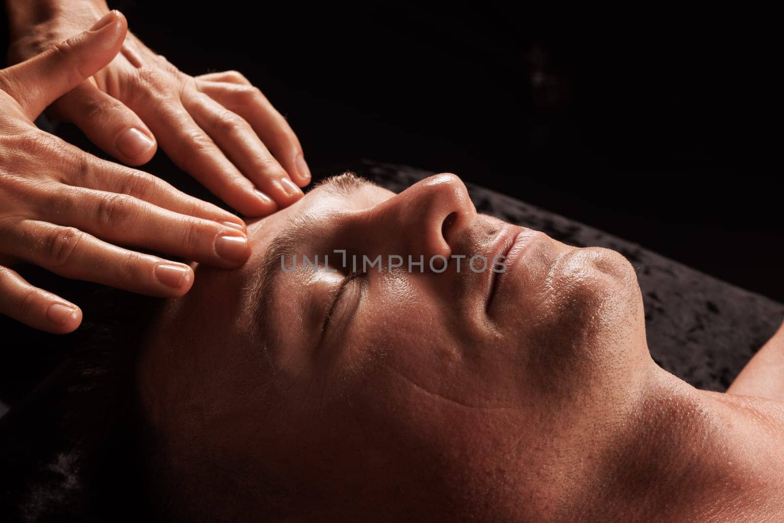 girl masseur doing facial massage to man on dark background close-up, facial skin rejuvenation, relaxation and meditation