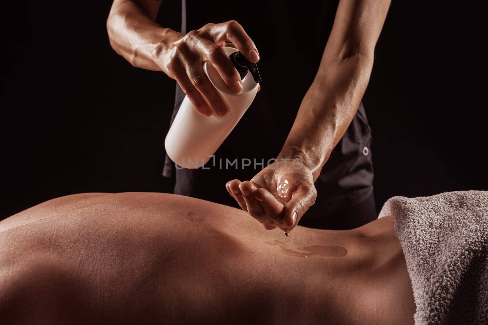 Girl giving back massage to muscular man on dark background close-up, pouring oil on body before massage