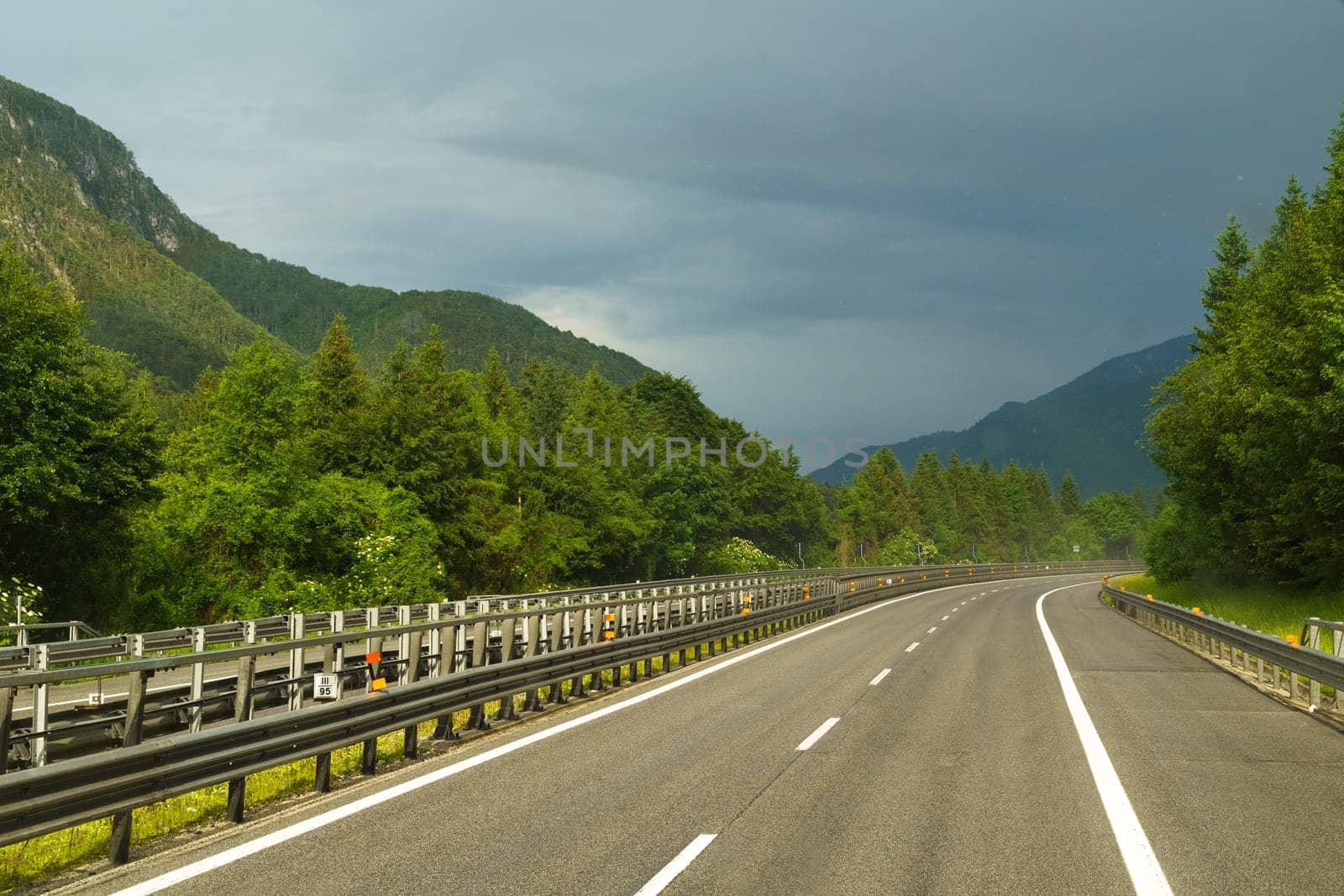 A narrow highway meanders through vibrant green trees, flanked by majestic mountains, as dark clouds gather ominously overhead in the afternoon.