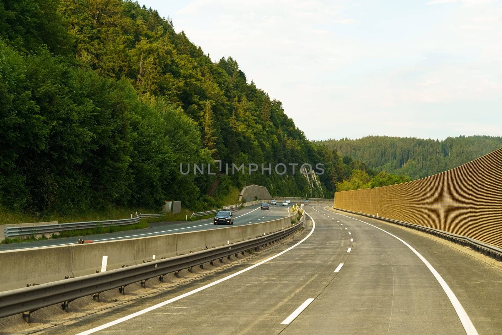 A serene highway weaves through verdant hills, as cars traverse the smooth asphalt under a gentle sunset glow, creating a picturesque evening journey through nature.