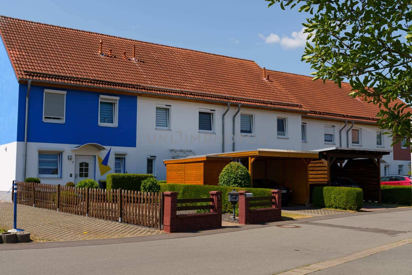 Waltershausen, Germany - June 11, 2023: A lovely residential structure in Waltershausen showcases a vibrant blue and white facade. The garden is neatly trimmed, with inviting hedges and shaded carports on a bright day.