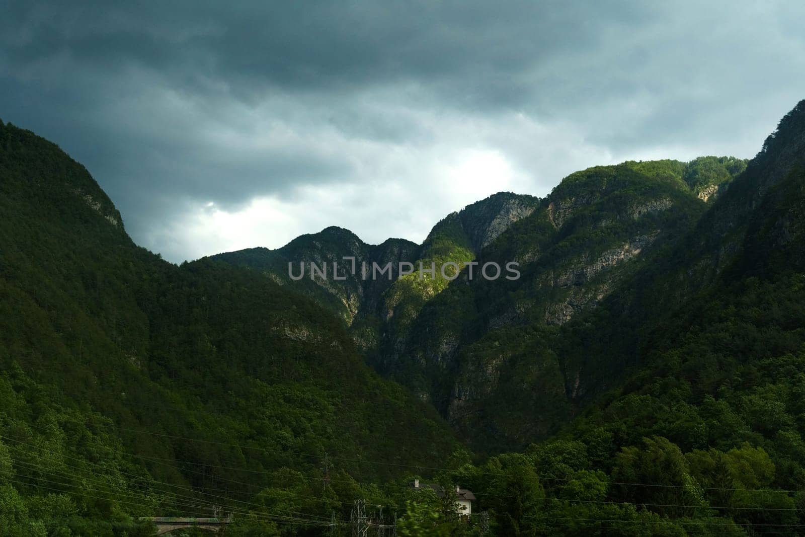 Dramatic cloud formations hover over towering mountains as the last light of day illuminates lush green valleys, creating a stunning contrast in the evening sky.