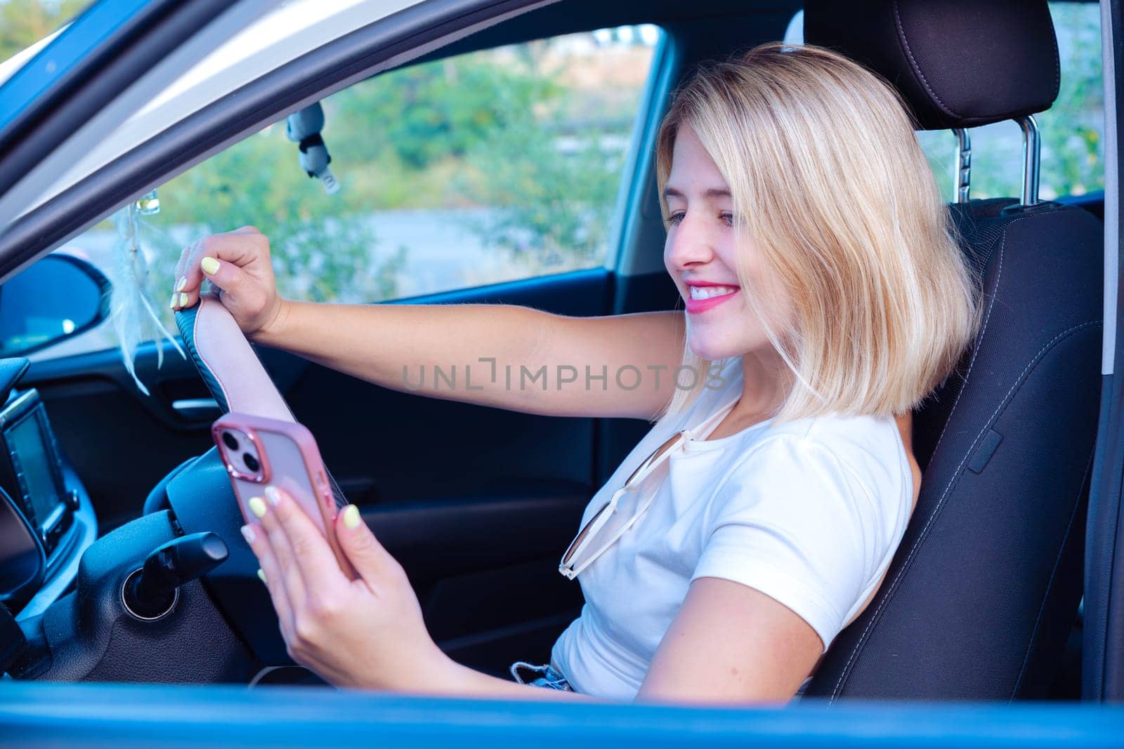 A young woman is sitting in the car holding her mobile phone, smiling with joy as she looks out a side window.