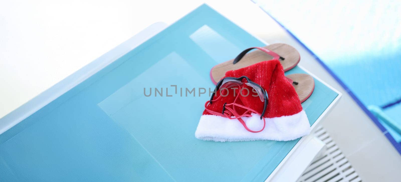 Hat of Santa Claus, swimming slippers on sunbed. Holidays near sea pool for Christmas holidays and relaxation, leisure, tourism concept.
