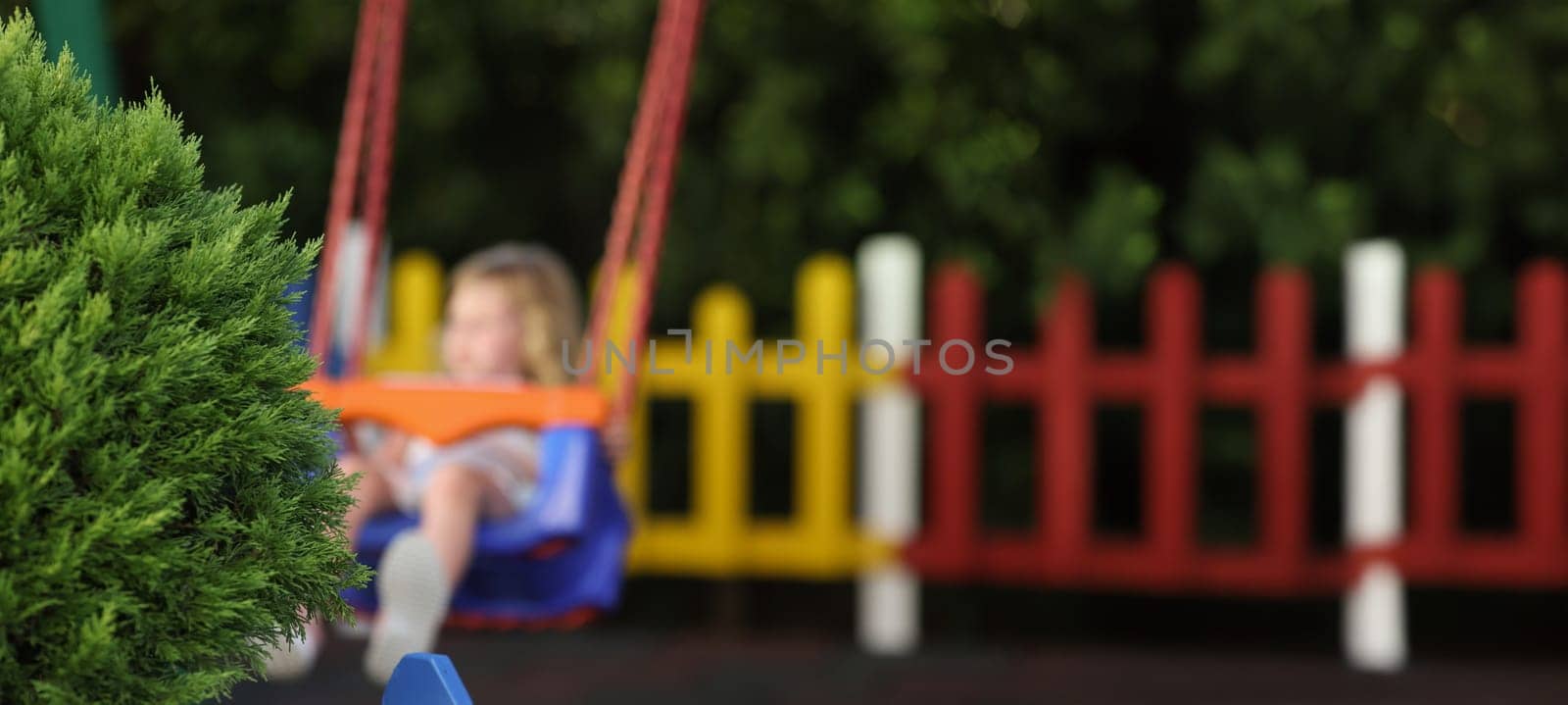 Close-up of green fluffy bush on playground child on swing in park alone. Little girl enjoy ride outdoors, place for children. Childhood, nature concept