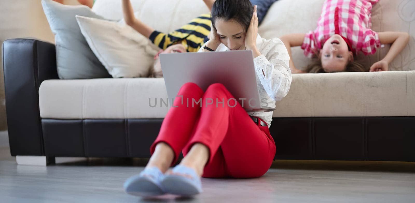 Low angle of mother try to concentrate and work while kids going crazy and jump. Female with pc close ears. Remote job, work from home, childhood concept