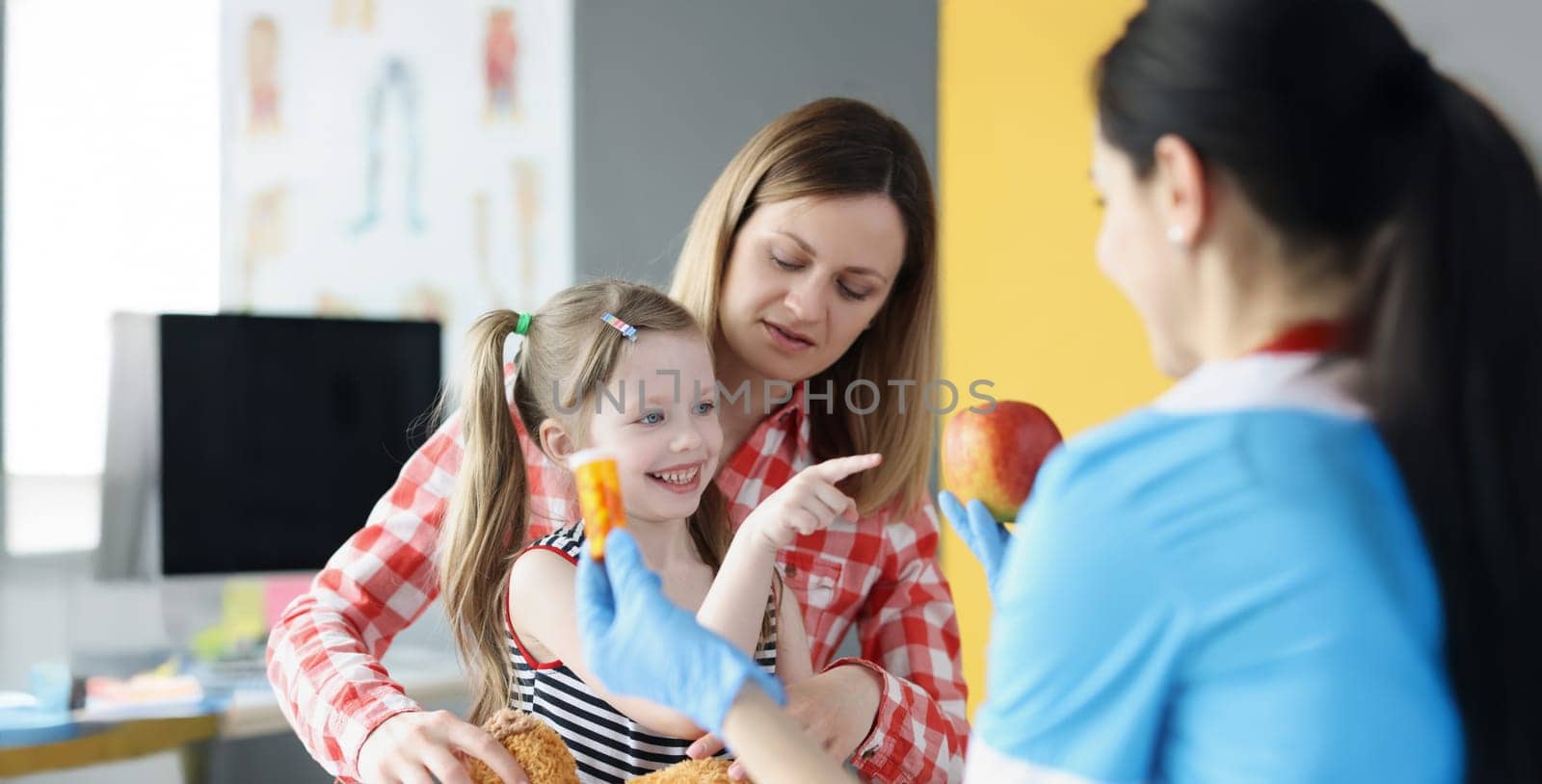 Portrait of happy little girl choose apple instead of bottle of medications. Medical worker in protective gloves and uniform. Pediatrician, clinic concept