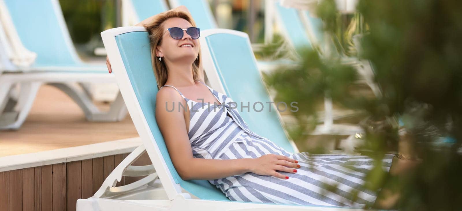 Portrait of happy young woman on sit on sunbed in pretty summer dress. Smiling relaxed female in sunglasses enjoy weather. Summer, chill, leisure concept