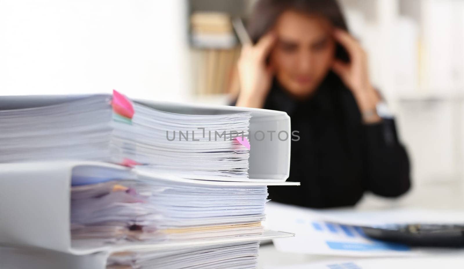 Close-up of tired and exhausted woman looks at documents touch head with hands. Huge pile of document folders on workplace. Headache, low energy concept