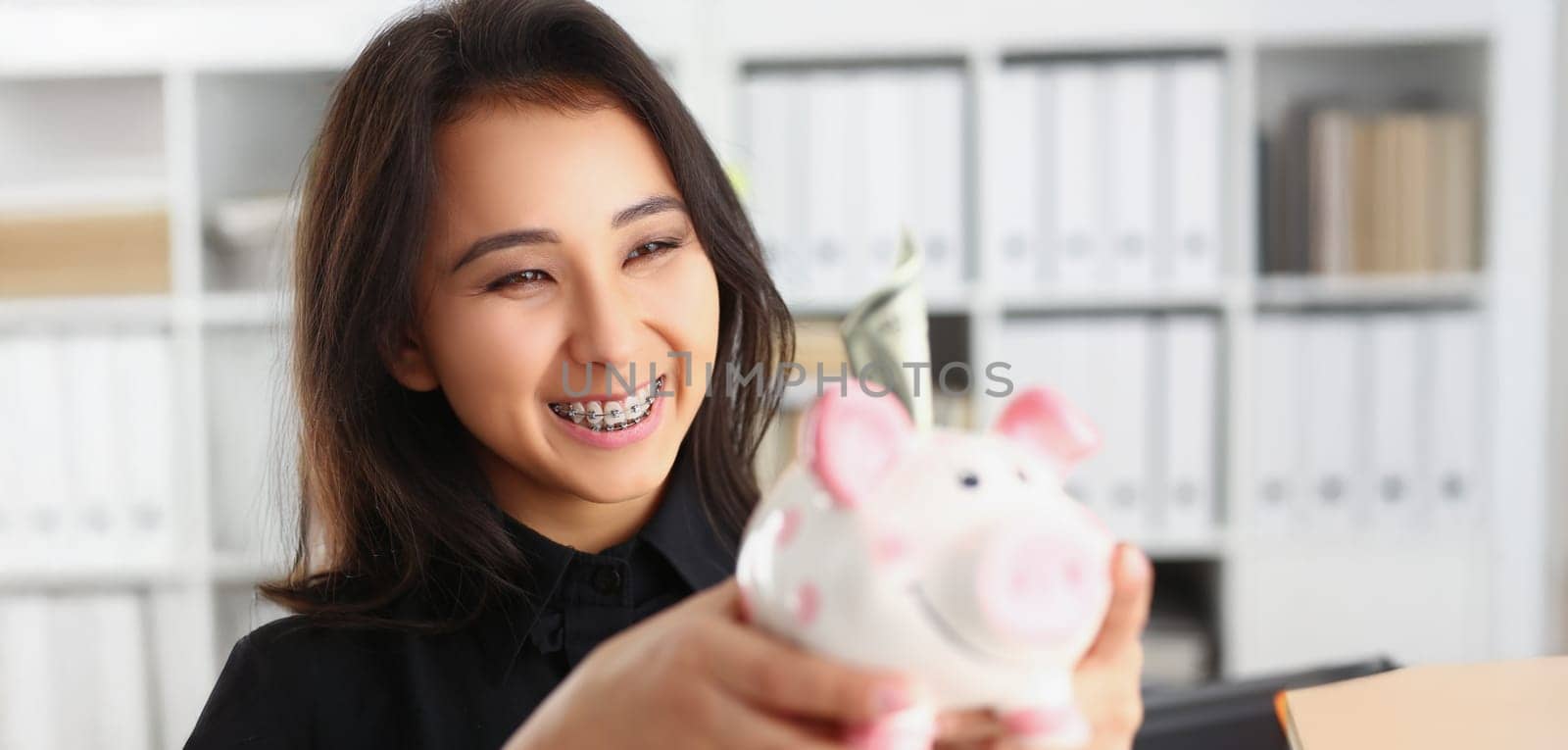 Portrait of pretty laughing young woman holding piggybank with banknote in it. Smiling brunette female save up for future. Investment, budget, fund concept