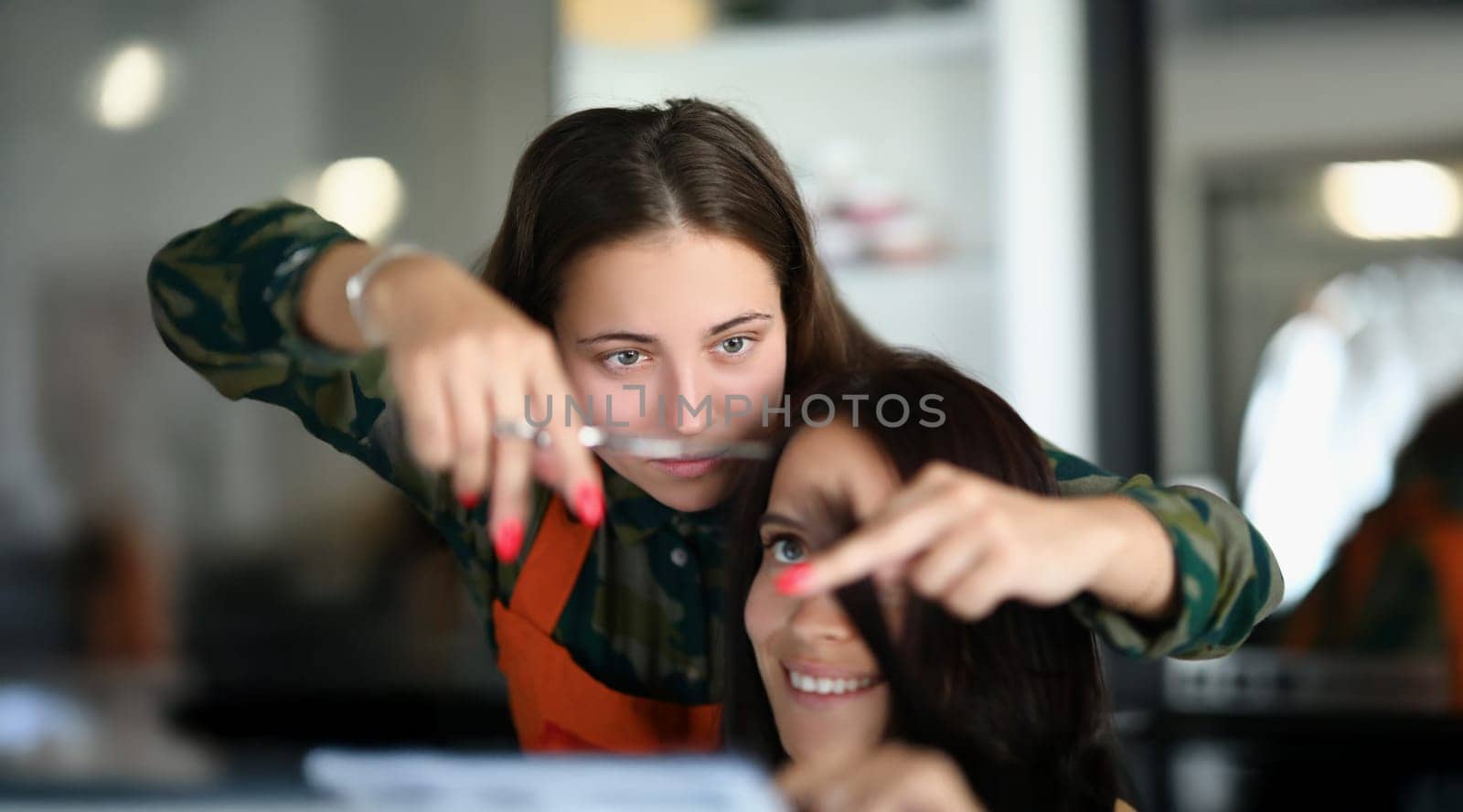 Portrait of concentrated professional hairdresser holding lock of hair and cutting with scissors. Happy female client sit in masters chair. Beauty concept